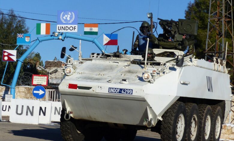 A white UN eight-wheeled armored vehicle marked UNDOF drives past a UN gate with multiple national flags and signage, in a Syrian Golan Heights setting under clear skies.