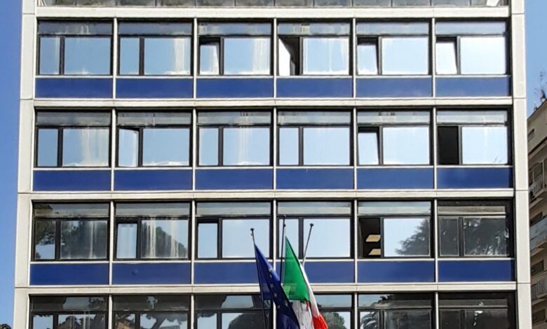 A frontal view of a modern mid-century office building with blue horizontal panels and many windows. Two flags—the Italian tricolour and the European Union flag—are mounted above the entrance.