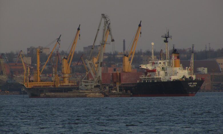 A seagoing cargo vessel berthed at Mykolaiv Sea Commercial Port, Ukraine, with multiple yellow and gray cranes and port infrastructure along the quay, photographed from across the water under overcast light.