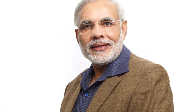 A studio portrait of an older man with a grey beard wearing glasses, a brown blazer, and a dark blue collared shirt, posed against a plain white background.
