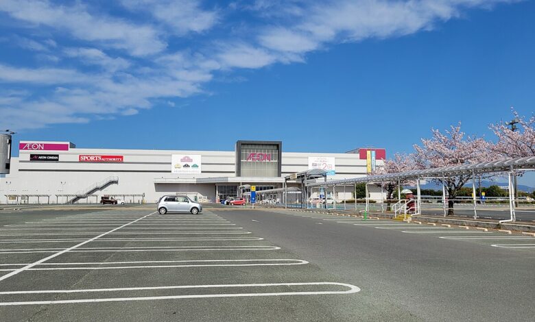 Wide exterior shot of AEON Mall Kumamoto with the mall’s facade and signage visible behind a large, mostly empty parking lot. Cherry blossom trees line parts of the lot under a blue sky with scattered clouds.