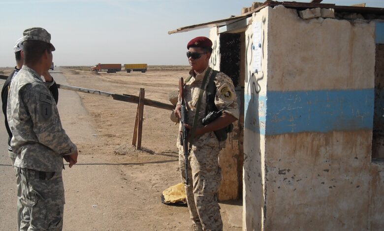 An Iraqi border policeman in desert camouflage with a red beret stands armed beside a small concrete checkpoint hut at a dusty border crossing, speaking with two U.S. uniformed personnel. Trucks and a flat, arid landscape extend into the background.