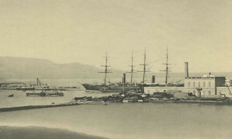 A historic sepia photograph of Damietta’s waterfront with several masted ships and small vessels moored by quays and a waterfront building, viewed across calm water.