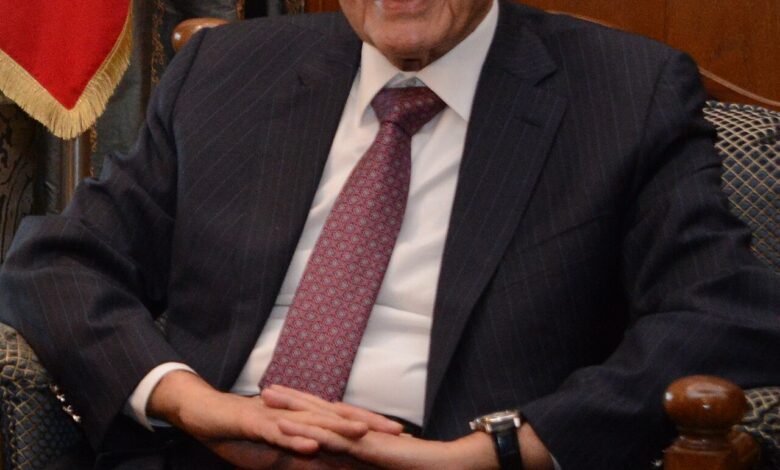 An older man in a dark pinstripe suit sits smiling in an ornate office setting; part of a Lebanese flag is visible beside him, with wood paneling and bookshelves in the background.