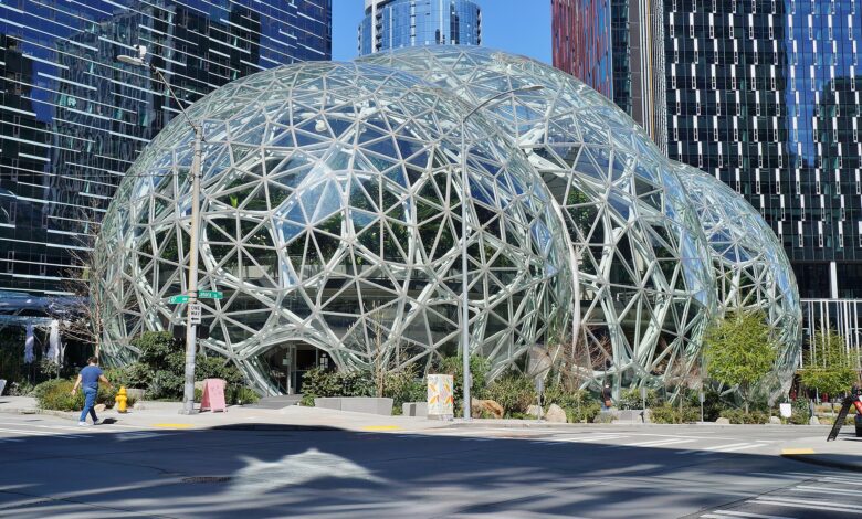 A clear street-level view of the glass-domed Amazon Spheres set among Amazon office towers in Seattle, with a few pedestrians and surrounding landscaping visible.