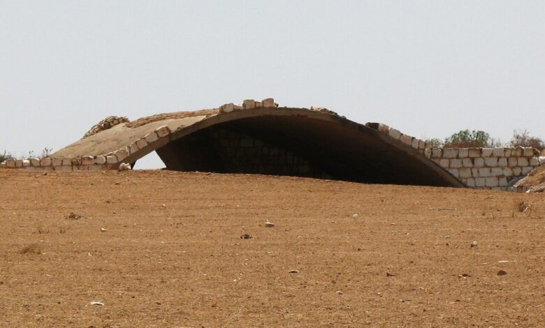 A derelict arched hardened aircraft shelter made of concrete and stone sits in a barren desert landscape under a clear sky.
