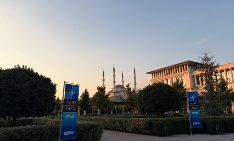 An exterior view of the Presidential Complex grounds in Ankara at sunset, with manicured hedges and trees in the foreground, a large mosque with four minarets in the background, and two event banners along the walkway.