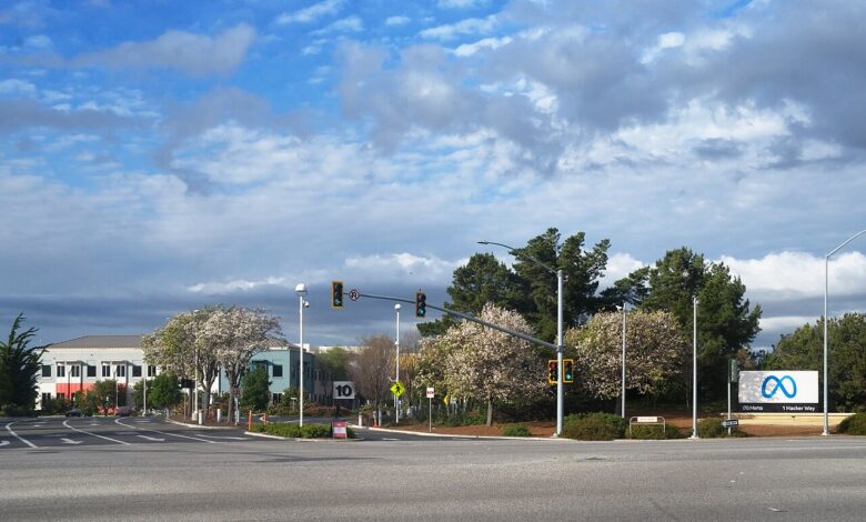 A wide view of the roadway and entrance area to Meta’s Menlo Park campus, with a roadside monument sign displaying the Meta infinity logo on the right, office buildings and flowering trees in the background under a blue sky with clouds.
