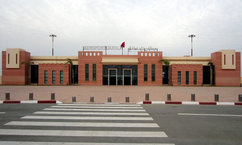 A frontal view of Dakhla Airport’s terminal building with the sign “AEROPORT DAKHLA” on the roof, a Moroccan flag, palm trees, and a pedestrian crossing in the foreground.