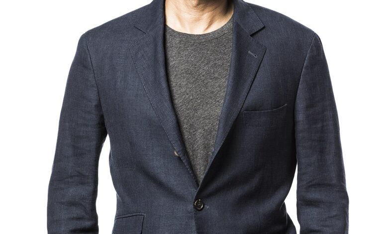 A smiling, bespectacled man in a dark blazer over a gray T‐shirt stands against a plain white background with hands in his pockets.