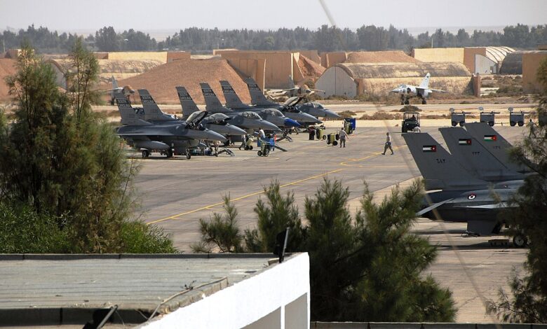 Daytime view of a busy military flight line with several F-16 fighter jets from Jordan and the United States parked on the tarmac at Muwaffaq Salti Air Base, with ground crews working and base structures and trees in the background.