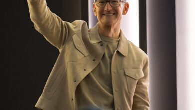 An older adult with short gray hair and glasses, wearing a beige jacket and light shirt, smiles and raises a hand in a wave inside a modern, softly lit indoor venue.