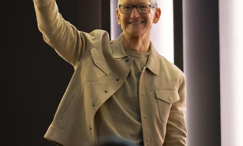 An older adult with short gray hair and glasses, wearing a beige jacket and light shirt, smiles and raises a hand in a wave inside a modern, softly lit indoor venue.