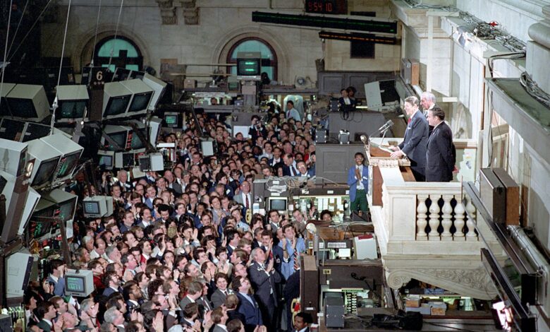 A wide view inside the New York Stock Exchange with a crowded trading floor full of people and monitors. On an interior balcony, several suited speakers address the assembled crowd, with trading screens and floor activity visible below.