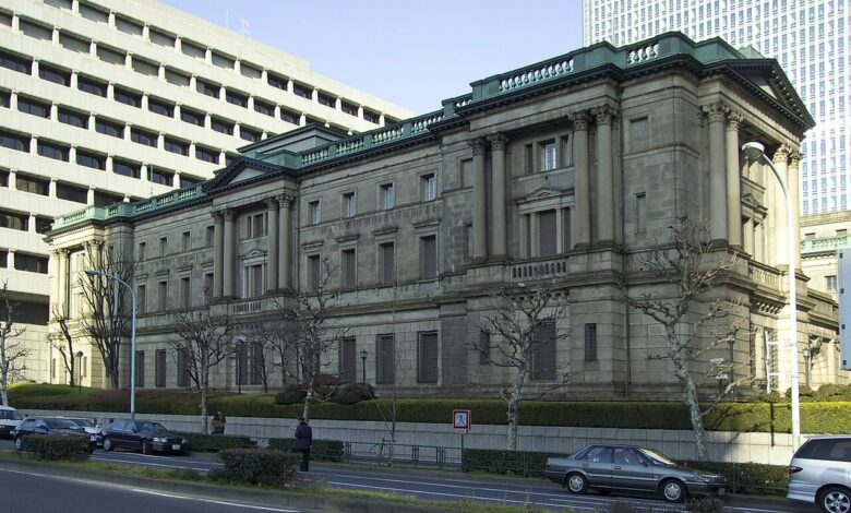 A street-level view of the historic stone Bank of Japan headquarters in Tokyo with its distinctive green-capped roof sections, flanked by modern office buildings and a roadway with parked cars in the foreground.