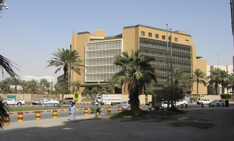 A tan, modern government building with horizontal brise-soleil panels and palm trees along a multi-lane street with passing cars in Riyadh under a clear sky.