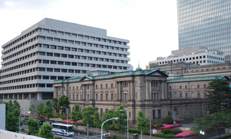 A wide view of the Bank of Japan head office in Tokyo, featuring the historic stone main building with green copper roof elements in the foreground, the modern annex building to the left, and a city street with vehicles and greenery in front.