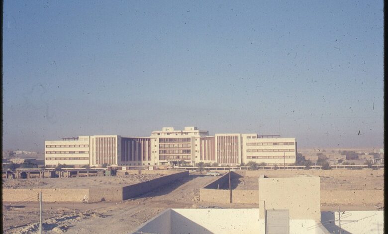 A wide, daylight view of a large, multi-wing government building set behind construction areas and low walls. The structure has a central block with lower wings extending to both sides, typical of mid-20th-century architecture, under a clear sky.