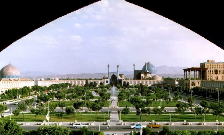 A wide, elevated view over Naqsh-e Jahan Square in Isfahan with manicured gardens, pathways, and surrounding arcades. The Shah Mosque dome and other historic buildings frame the square under a bright sky.