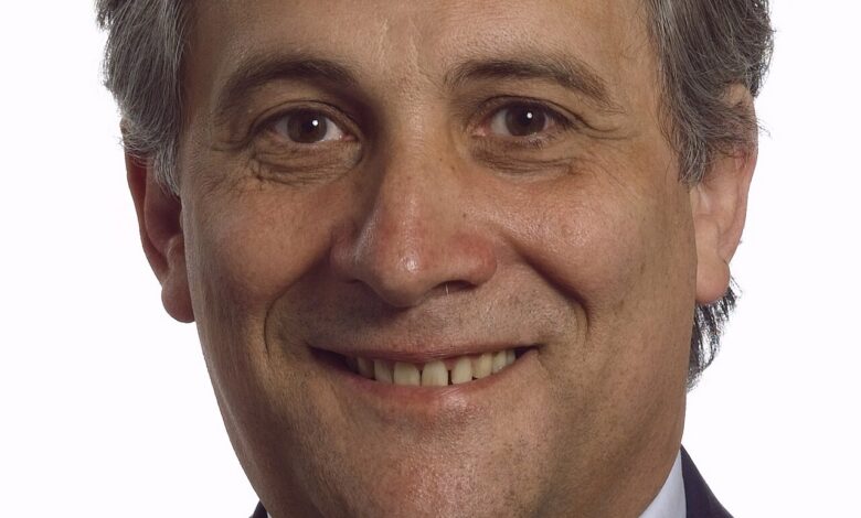 A formal head-and-shoulders studio portrait of a middle‐aged man in a dark suit, light blue shirt, and patterned tie against a plain white background, smiling toward the camera.