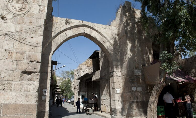 A wide street scene in Aleppo showing the historic Bab al‐Maqam stone gate under a clear blue sky. Pedestrians and a pushcart are visible near shops along the cobbled street.