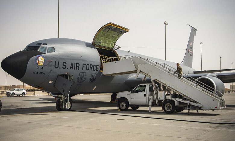 A U.S. Air Force KC-135 Stratotanker is parked on a concrete flight line with its forward cargo door open and mobile stairs positioned at the side. Light poles, base buildings, and equipment are visible in the background under a hazy sky.