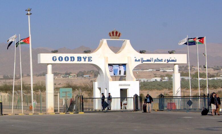 The Jordanian exit gate at the Aqaba–Arava border crossing, with large “GOOD BYE” sign, Jordanian flags, a portrait on a screen, and travelers near the gate against a desert mountain backdrop.