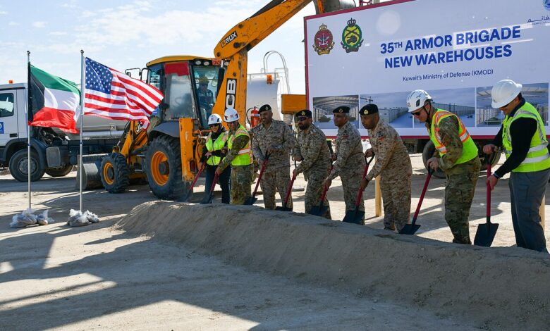 A line of uniformed personnel and construction staff in safety vests and helmets shovel sand at a groundbreaking ceremony. Behind them are excavators, a large project sign reading “35th Armor Brigade New Warehouses” with Kuwait’s Ministry of Defense mentioned, and Kuwaiti and U.S. flags.