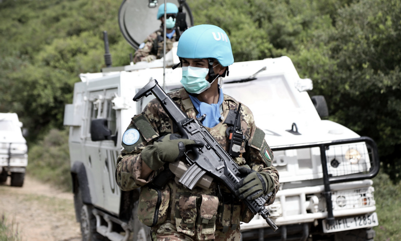 UN peacekeepers in blue helmets and masks, armed and patrolling on a dirt track beside white armored vehicles marked UN in a green, hilly area.
