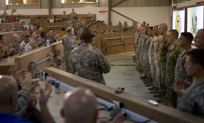 U.S. service members in uniform inside a large briefing room with tiered seating. A senior officer stands at the front addressing a line of troops while others seated nearby applaud.