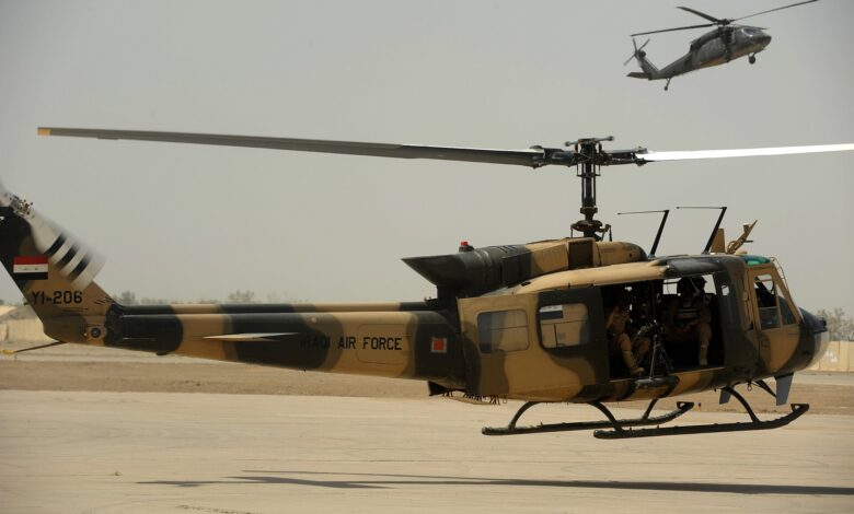 An Iraqi Air Force UH-1H Huey helicopter with "Iraqi Air Force" markings lifts off from a runway at Taji Air Base, Iraq, with another helicopter flying in the background under a clear sky.