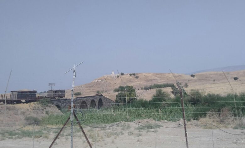 A dry valley landscape with a wire border fence in the foreground and the old multi-arch Gesher bridge structure and hills beyond, marking the Israel–Jordan border area.
