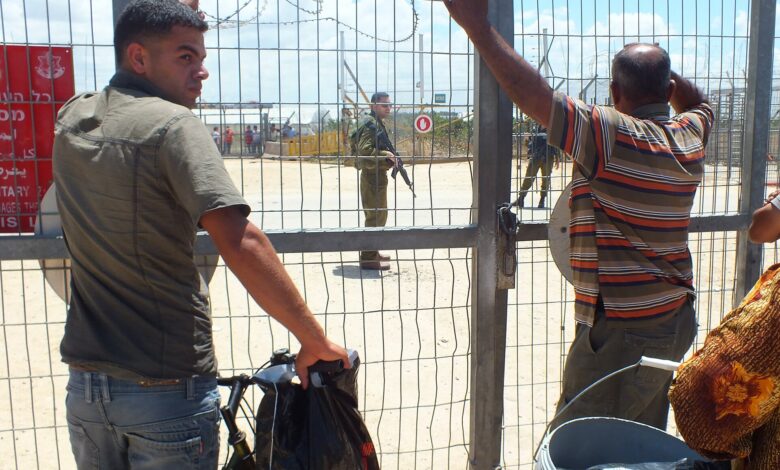 At a fenced agricultural gate in the West Bank, several Palestinian civilians wait at the barrier with bicycles and bags while two Israeli soldiers stand on the other side. The scene includes wire fencing, barbed wire, warning signs, and a dusty road under daylight skies.