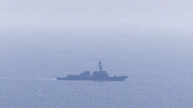 A distant view of a U.S. Navy guided-missile destroyer underway on a hazy, sunlit sea, with a faint silhouette of another vessel on the horizon.