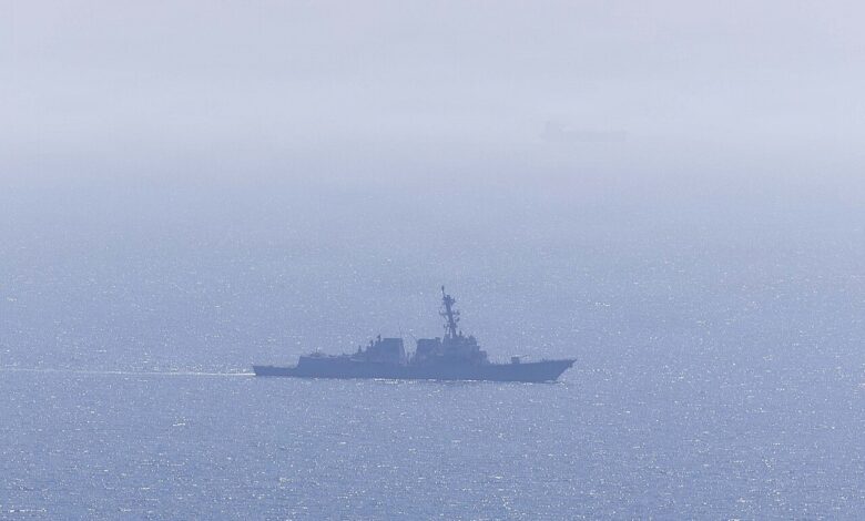 A distant view of a U.S. Navy guided-missile destroyer underway on a hazy, sunlit sea, with a faint silhouette of another vessel on the horizon.