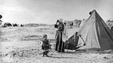 A black‐and‐white scene of a refugee camp in Khan Younis with canvas tents pitched on sandy ground. A woman stands beside a tent with a small child nearby; additional tents and sparse vegetation are visible in the background under an open sky.