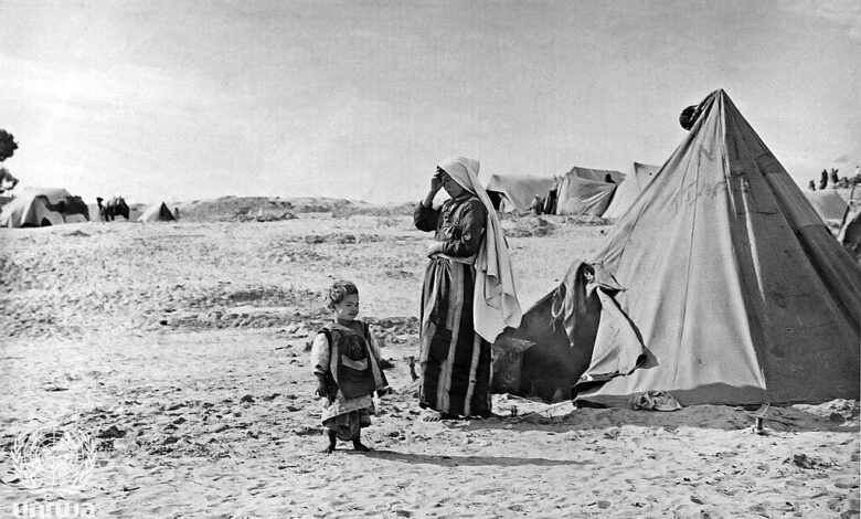 A black‐and‐white scene of a refugee camp in Khan Younis with canvas tents pitched on sandy ground. A woman stands beside a tent with a small child nearby; additional tents and sparse vegetation are visible in the background under an open sky.