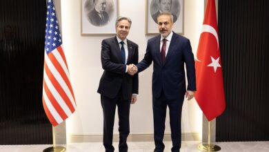 Two senior officials stand side by side for a handshake in an official interior, flanked by the U.S. and Turkish flags, with framed portraits on the wall behind them.