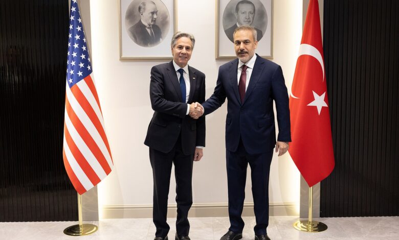 Two senior officials stand side by side for a handshake in an official interior, flanked by the U.S. and Turkish flags, with framed portraits on the wall behind them.