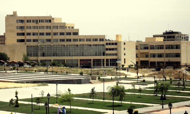 A wide view of the Aleppo University Faculty of Arts and Humanities complex with beige modernist buildings and landscaped grounds in the foreground under a bright sky.
