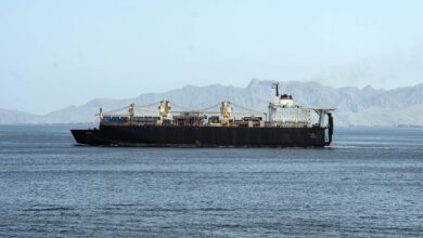 A cargo ship sails across calm blue waters in the Strait of Hormuz with a rugged, mountainous shoreline in the background under a clear sky.