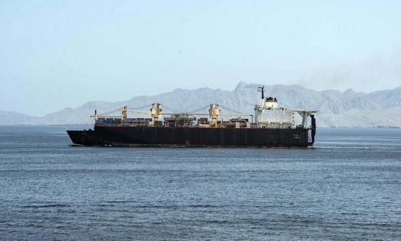 A cargo ship sails across calm blue waters in the Strait of Hormuz with a rugged, mountainous shoreline in the background under a clear sky.