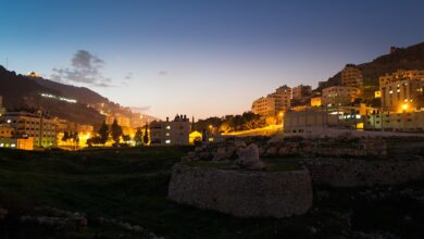 A dusk cityscape of Nablus with hillside neighborhoods lit by streetlights. In the foreground are low archaeological stone walls and grassy ground; in the background, buildings climb the surrounding hills under a darkening sky.