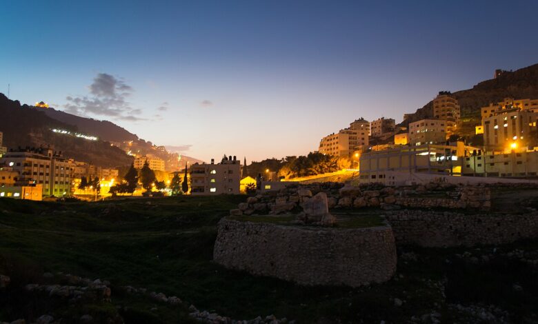 A dusk cityscape of Nablus with hillside neighborhoods lit by streetlights. In the foreground are low archaeological stone walls and grassy ground; in the background, buildings climb the surrounding hills under a darkening sky.