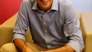 A man in a blue striped shirt with rolled-up sleeves sits on a beige sofa, smiling with hands clasped. A red panel with partial text is in the background and sheer curtains at right.