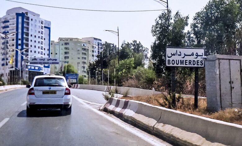 A roadway entering an urban area with mid-rise apartment buildings; a clear roadside sign reads “BOUMERDES” in Arabic and Latin script. Vehicles are driving on a divided road lined with trees and streetlights.